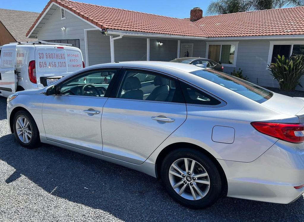 Silver car parked in front of a house with a white van in the background.